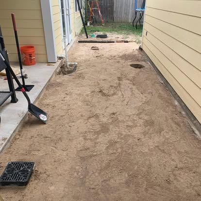 A yard with a patch of cleared, sandy soil between yellow siding and a concrete walkway, featuring tools and a drain grate.