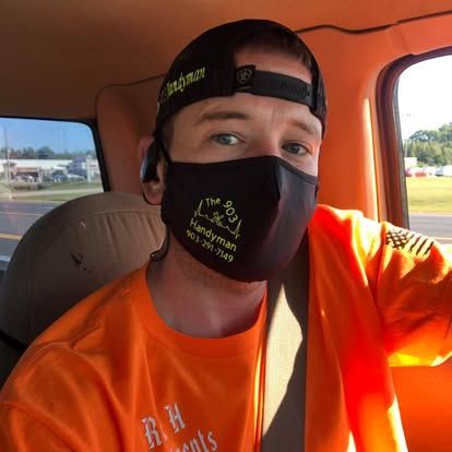 A person in an orange work shirt and black mask with branded text sitting in the driver's seat of a vehicle.