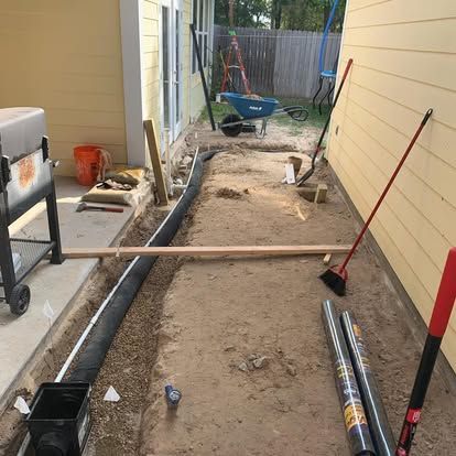A drainage pipe is being installed in a trench along the side of a yellow house, with tools and construction materials nearby.