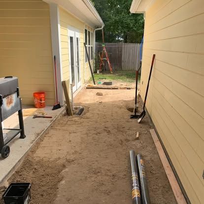 An outdoor side yard showing a dirt path between two yellow walls, with tools, building supplies, and a grill nearby.