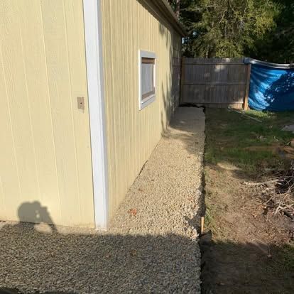 A light-yellow shed wall with a small window, alongside a freshly laid gravel path leading to a backyard fence.