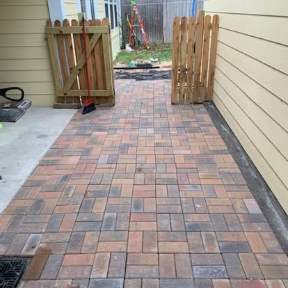 A pathway of multi-colored, rectangular paving stones leading to a wooden gate between two light yellow house walls.