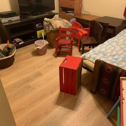 A cluttered room with a bed, a television stand, several small chairs, and a red wooden crate on the floor.