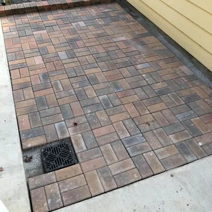A ground-level view of a multi-colored brick patio bordered by a concrete walkway, yellow siding, and a black storm drain.