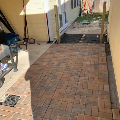 A newly installed herringbone-patterned brick paver patio between yellow building walls, leading to an open backyard area.