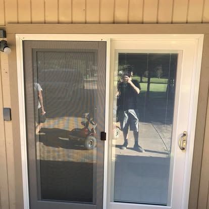 A person standing outside holds a phone to take a selfie reflected in a glass sliding patio door with a screen panel.