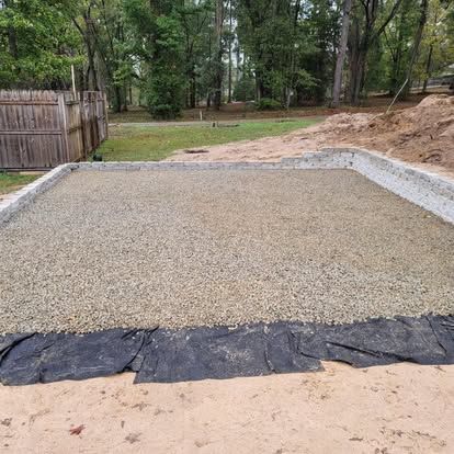 A prepared rectangular foundation area filled with gravel, bordered by light-colored blocks and black landscaping fabric.