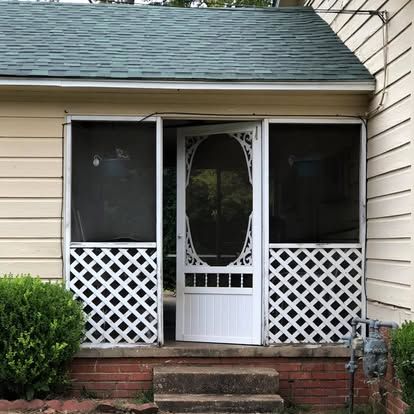 A yellow-sided house porch with a white lattice base, a central decorative screen door, and a green shingled roof.