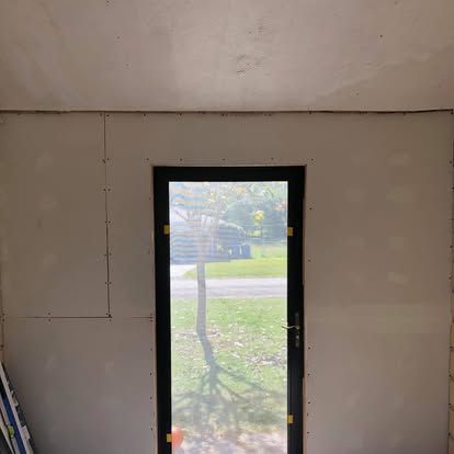 Interior view of a door frame set into a freshly installed drywall wall, looking out at a grassy yard.