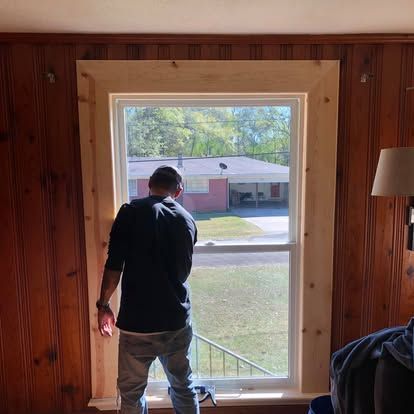 A person in dark clothing installs new light-colored wood trim around a window in a room with dark wood paneling.