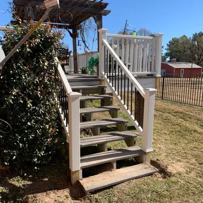 A set of wooden stairs with white railings and black balusters leads up to a deck in a backyard on a sunny day.