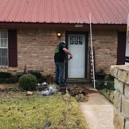 A person stands by the front door of a brick home with a red metal roof, appearing to work on the entryway landscaping.