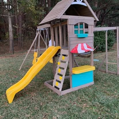 Wooden playset in a grassy yard featuring a slide, a ladder, swings, and a small play counter with a striped canopy.