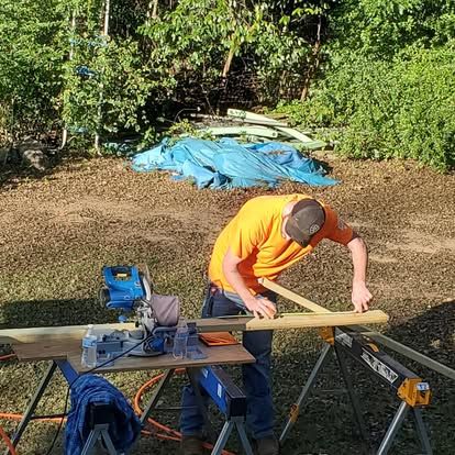 A person in an orange shirt uses a miter saw on a wooden board atop a workbench in an outdoor area.