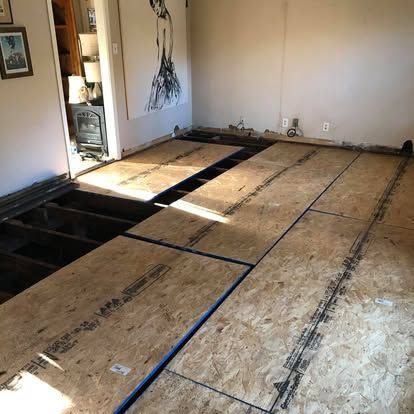 An indoor room undergoing renovation, showing exposed floor joists and newly installed plywood subflooring.