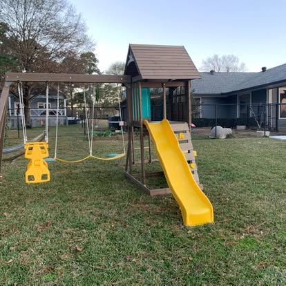 A brown wooden outdoor swing set with a yellow slide and a yellow baby swing, positioned in a grassy backyard.