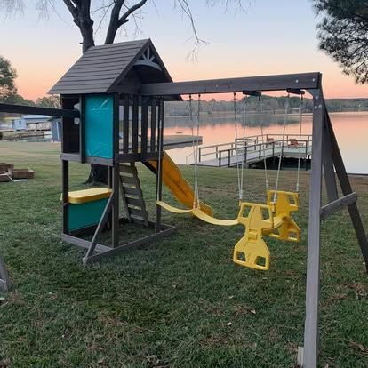 A wooden playset with a yellow slide and swings stands in a grassy yard overlooking a calm lake at sunset.