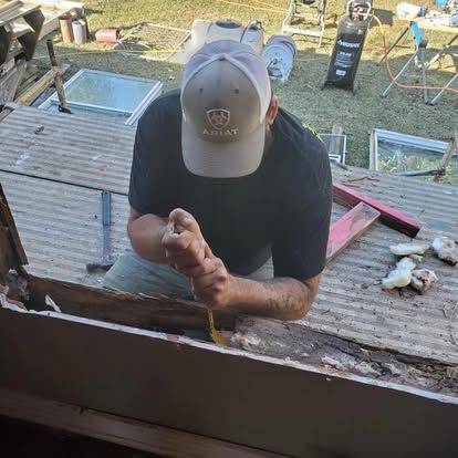 A person in an Ariat cap uses a pry bar to remove rotted wood from a structure during a home renovation project.