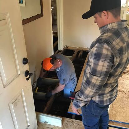 A contractor in an orange cap works on exposed subflooring while a supervisor in a plaid shirt observes nearby.