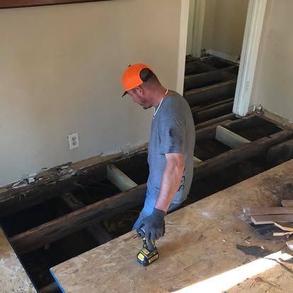 A worker wearing a bright orange hat stands over an open floor, using a drill on exposed wooden floor joists.