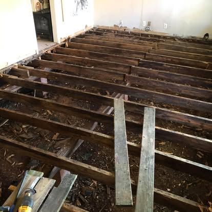 An indoor renovation scene showing exposed wooden floor joists over a dirt crawl space, with tools resting in the foreground.