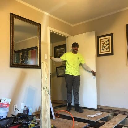 A person in a neon yellow shirt stands in a doorway during a room renovation, smiling with the floor under construction.