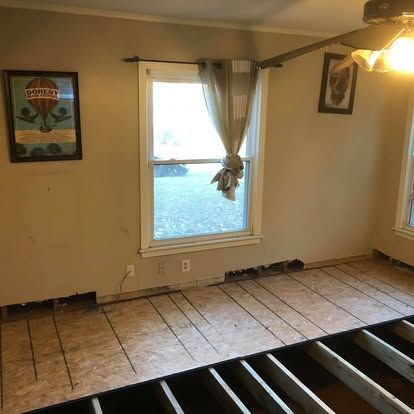 A room under renovation with exposed floor joists, partial plywood subflooring, beige walls, and a window with a sheer drape.