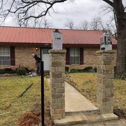 A person works in the yard of a brick home with a red metal roof, framed by two stone entrance pillars holding light boxes.