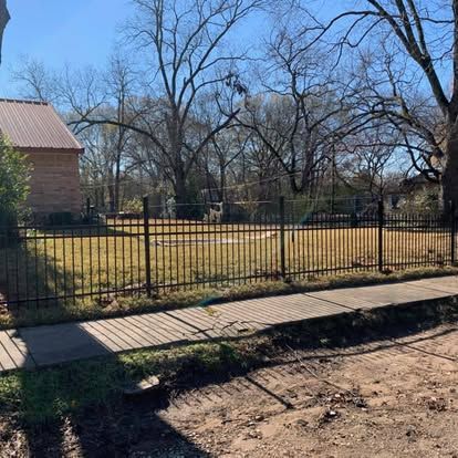 A black metal fence surrounds a residential lawn under a clear blue sky, next to a brick house and a sidewalk.
