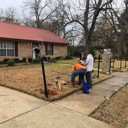 Two workers install a black metal fence in a residential front yard with a brick house.