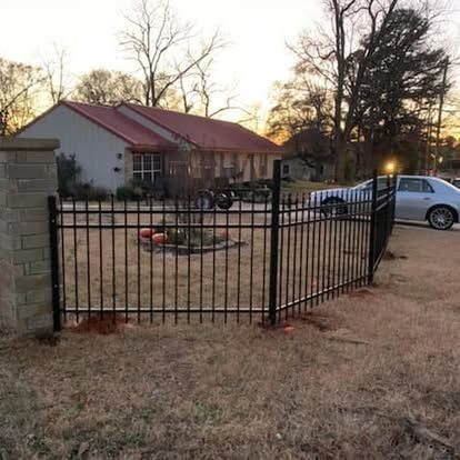 A black metal fence encloses a front yard in front of a house with a red roof, with a silver car parked in the driveway.