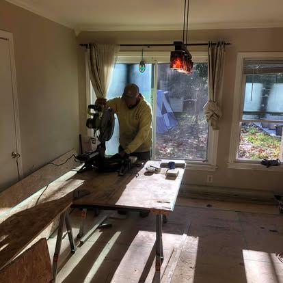 A person in a yellow hoodie cuts a piece of wood on a miter saw inside a room with a large window and bare flooring.