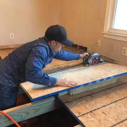 A person in a blue shirt and black cap uses a circular saw to cut a plywood subfloor in a room under construction.