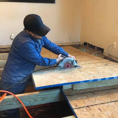 A person wearing a blue shirt and black cap uses a circular saw to cut a sheet of plywood subflooring in a room.