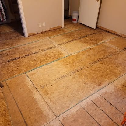 A room with a subfloor made of large, light brown OSB panels installed over floor joists, with a white door in the back.