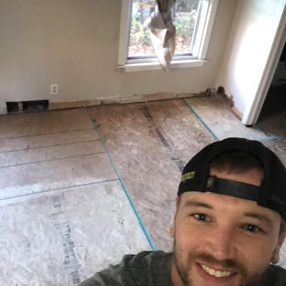 A smiling person in a black cap in an unfurnished room with exposed plywood subflooring and a window.