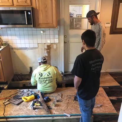 Three people wearing work clothes work on a kitchen renovation, installing new subflooring near a door.