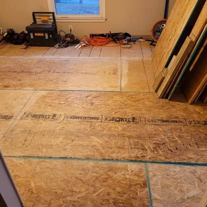 Subfloor plywood sheets installed on a room floor, with construction tools and stacked wood panels in the background.