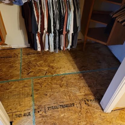 A closet with clothes hanging on a rack above a newly installed subfloor made of oriented strand board panels.