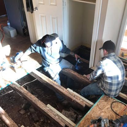 Two construction workers kneel on wooden floor joists, installing new subflooring in an unfinished room.