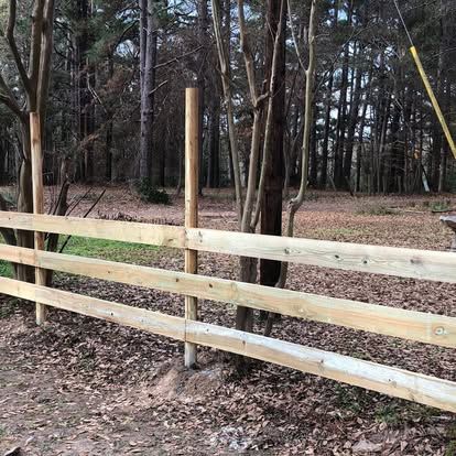 A three-rail wooden fence stands in a wooded area with fallen leaves on the ground.