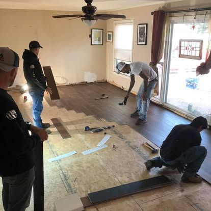 Three people working on installing dark wood plank flooring in a living room with a ceiling fan and large window.