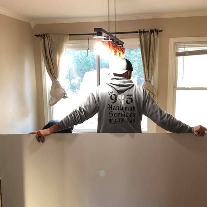A person wearing a grey branded hoodie stands in a room holding a large, white drywall sheet in front of them.