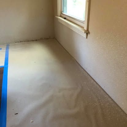 A room with beige walls and a window, the floor covered in protective brown paper and blue painter's tape.