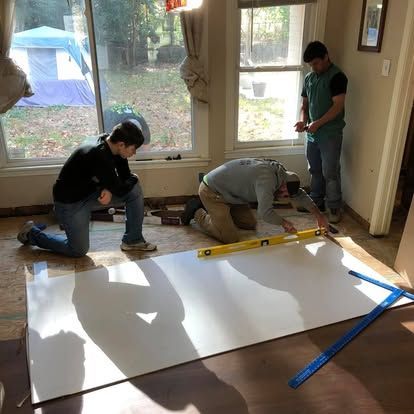 Three people work together on a home renovation project, measuring and leveling a large white panel on a wooden floor.