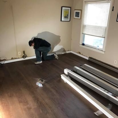 A person kneels to install dark wood-look flooring in a room with beige walls and a window with blinds.