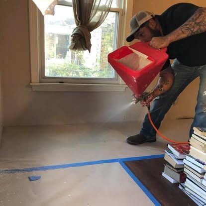 A person uses a red hopper spray gun to apply texture to a room wall with taped floors near a window.