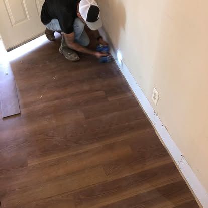 A person wearing a hat and black shirt kneeling on wood-look flooring to install white baseboard molding along a wall.