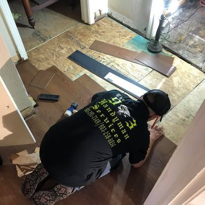A person wearing a branded black shirt kneels while installing dark brown laminate flooring in a room.