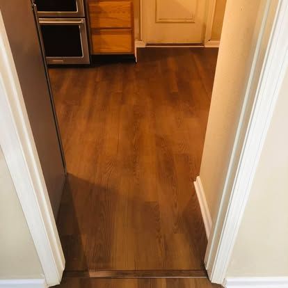 A view through a doorway into a kitchen with wood-look flooring and light wood cabinets, separated by a metal floor trim.